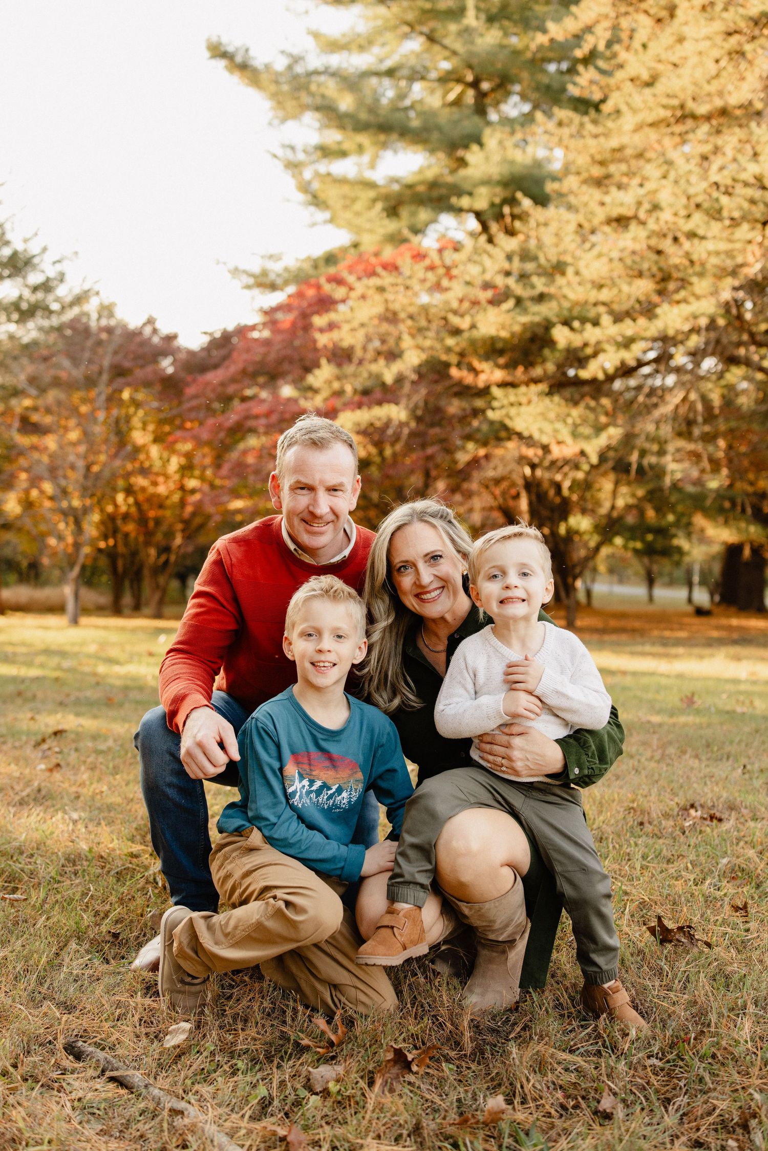 Family of four smiling together outdoors in a park during autumn, surrounded by colorful fall foliage.
