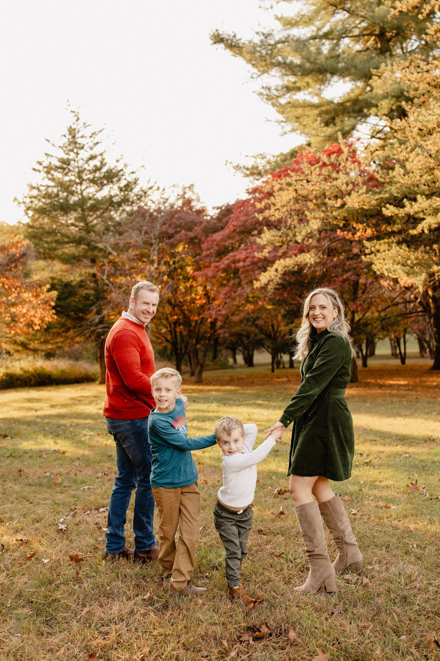 Family of four posing outdoors in fall foliage, wearing colorful autumn outfits, smiling in a park with vibrant red and gold trees.