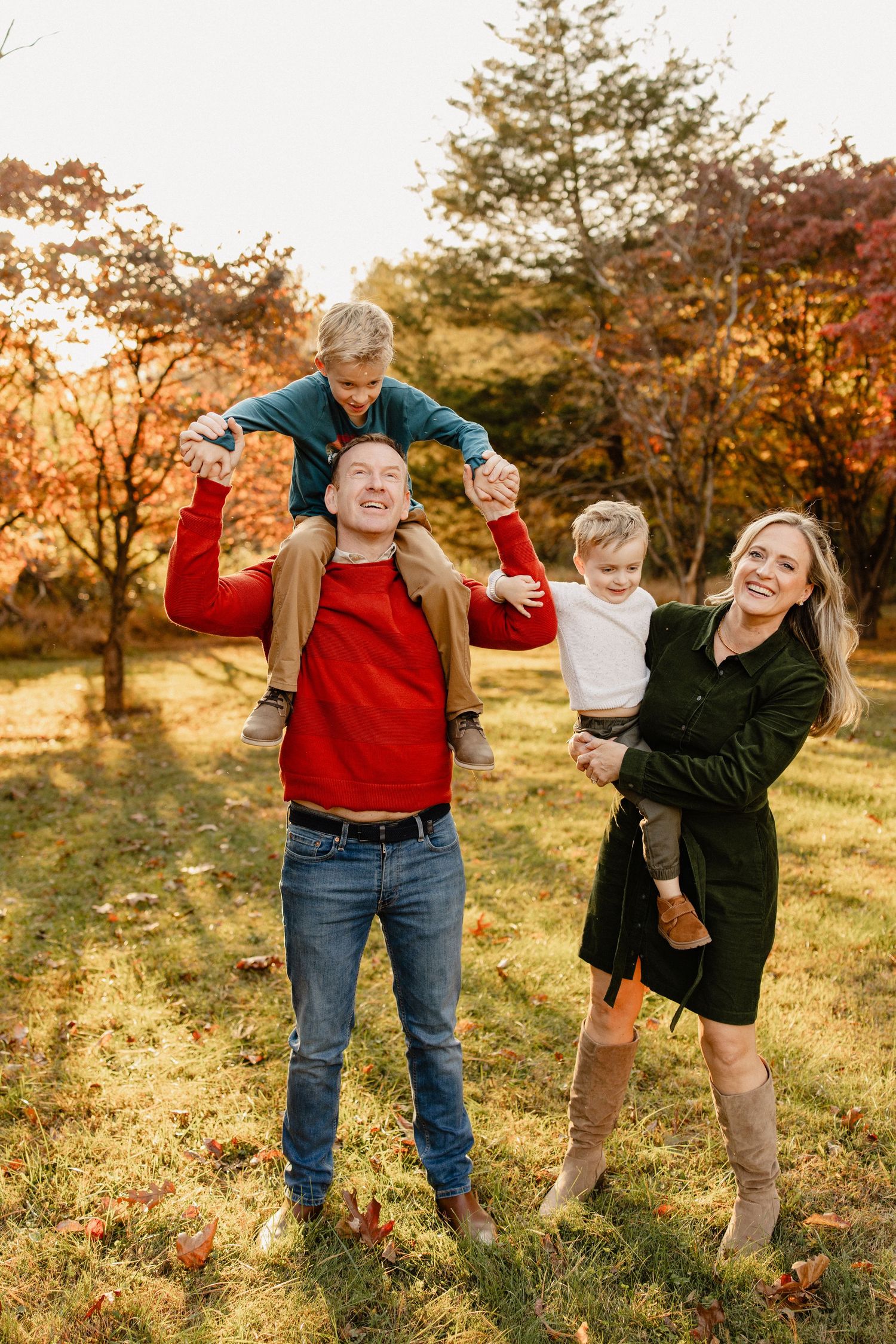 Family of four enjoying autumn outdoors, dad carries son on shoulders while mom holds toddler, surrounded by fall foliage.