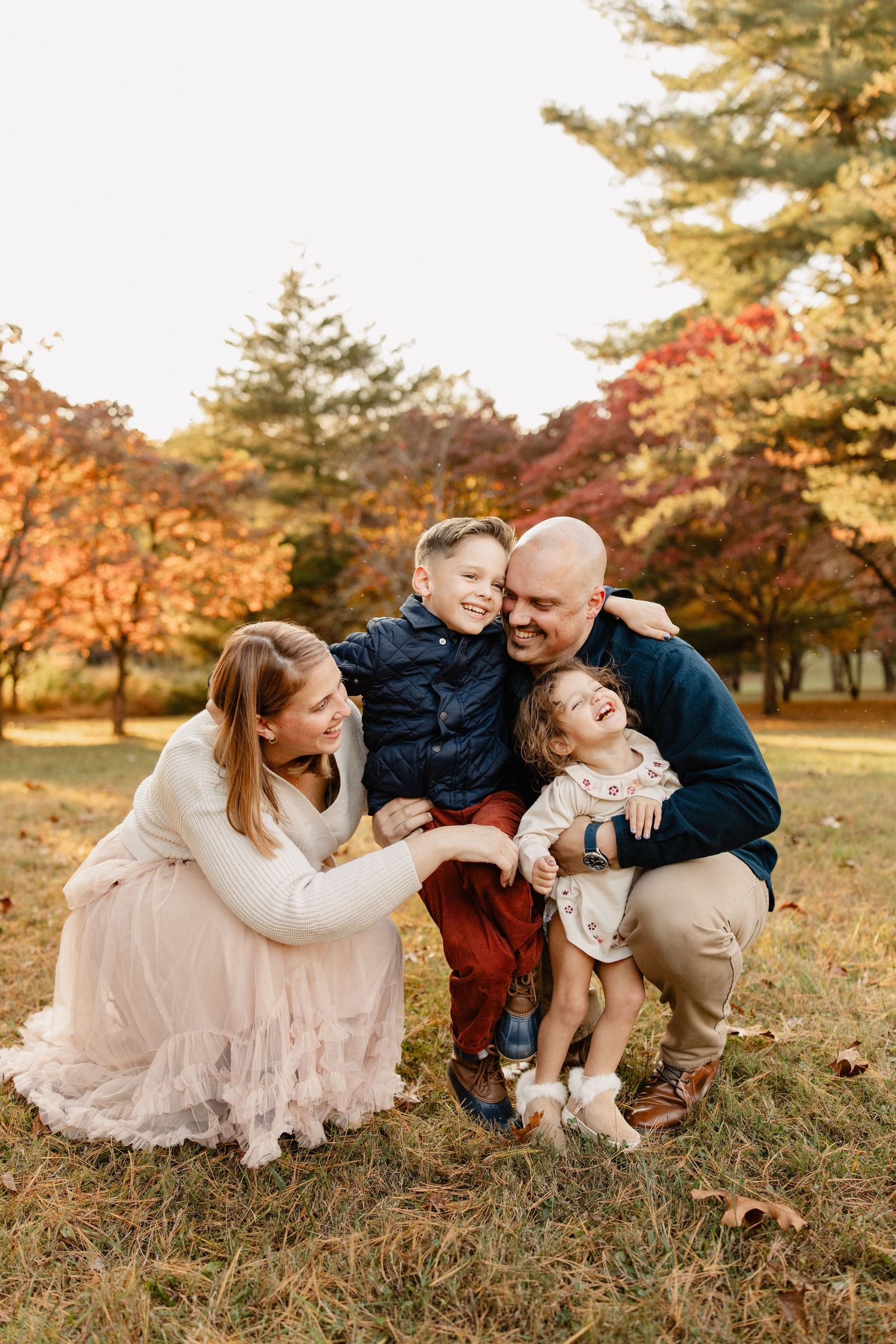 Family of four laughing together outdoors during autumn, parents crouching with two young children among colorful fall foliage.