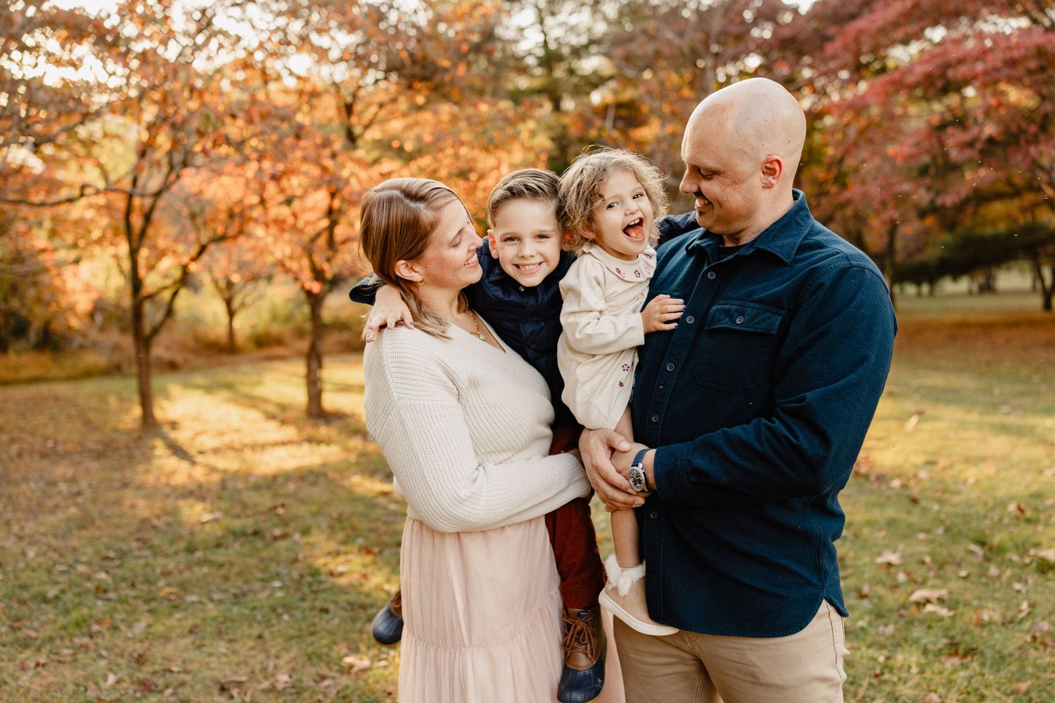 Happy family of four laughing together outdoors in autumn park with colorful fall foliage in background.