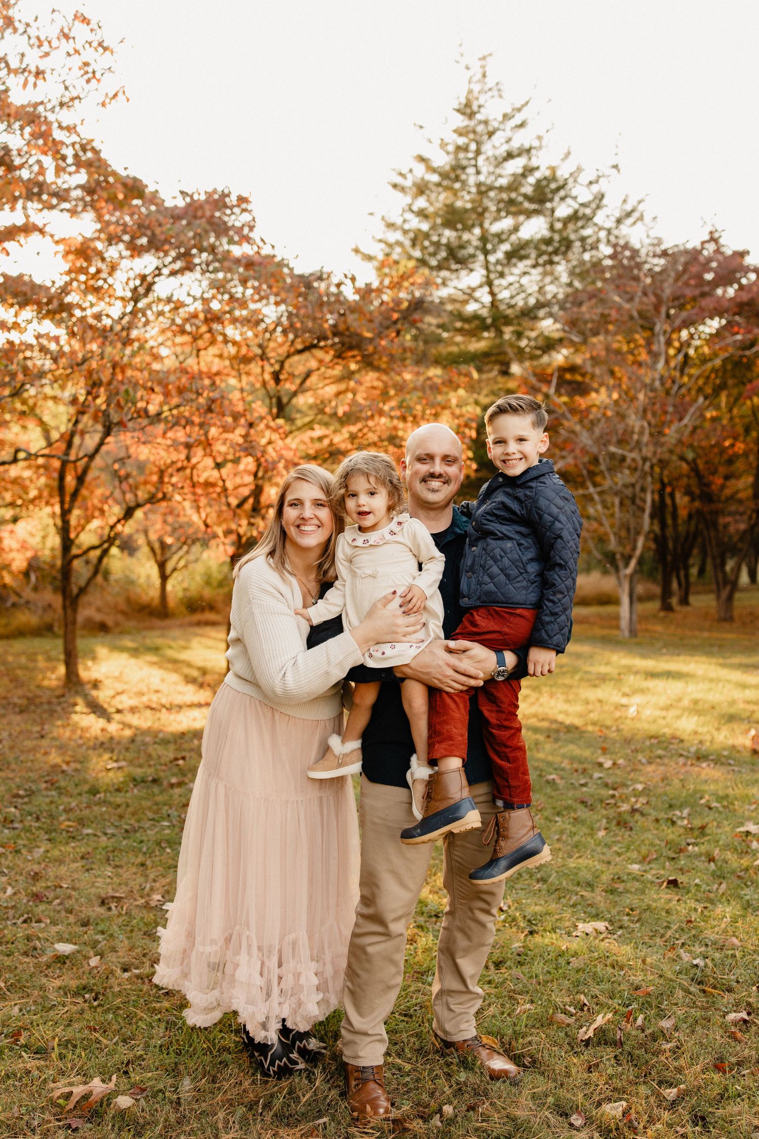 Family of four posing outdoors among vibrant autumn foliage, parents holding two young children while smiling.