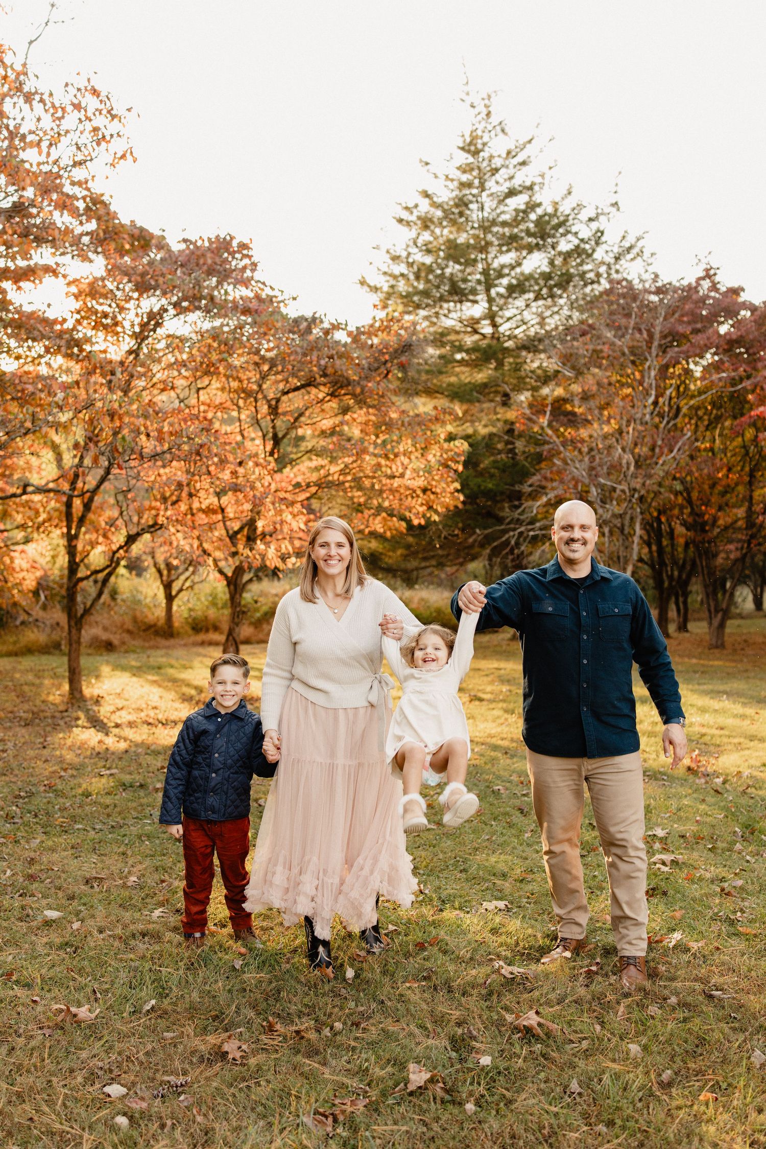 Family of four posing outdoors in autumn park, parents swinging toddler girl while young boy stands beside them.