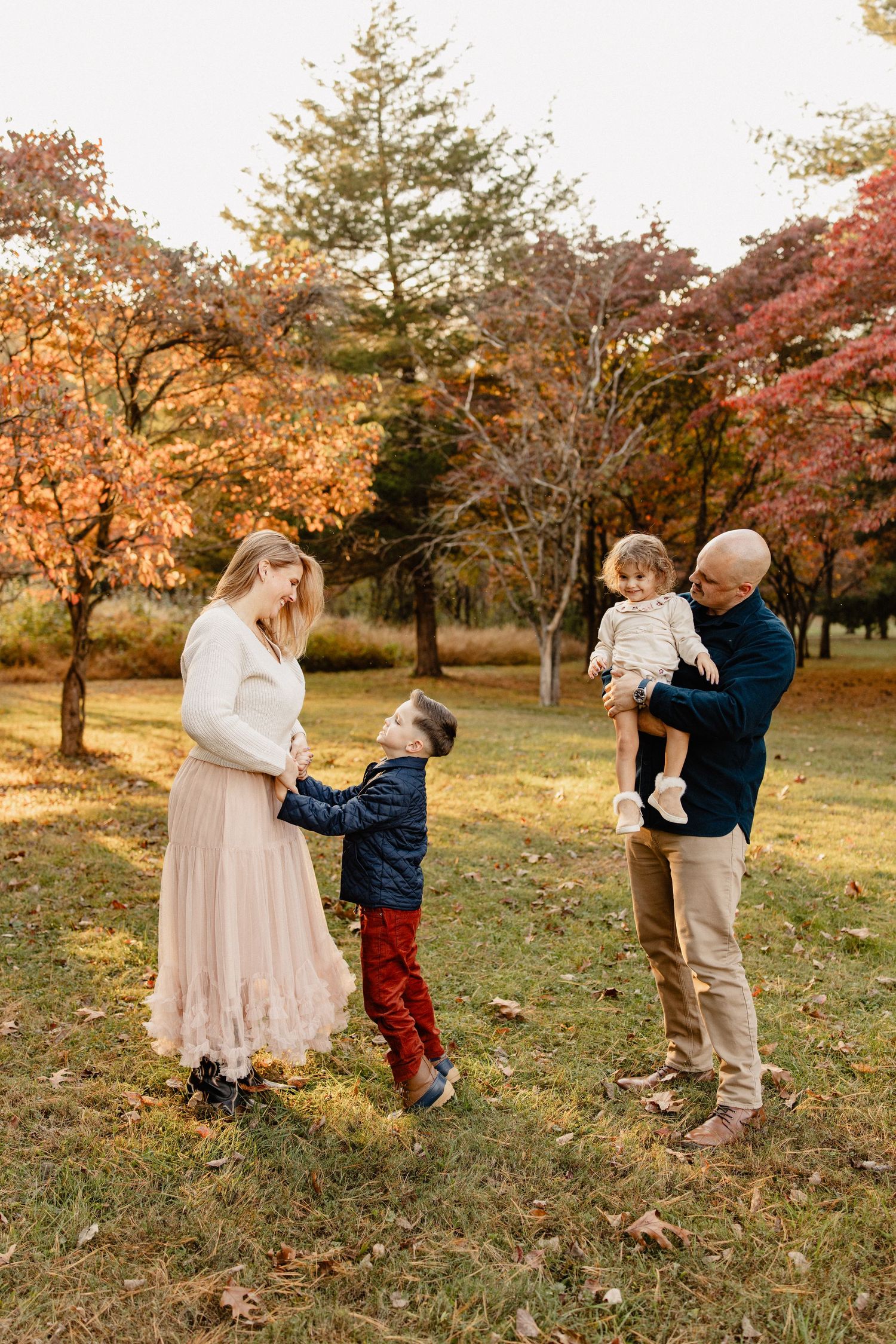 Family of four enjoys autumn park, parents holding children among vibrant fall foliage trees.