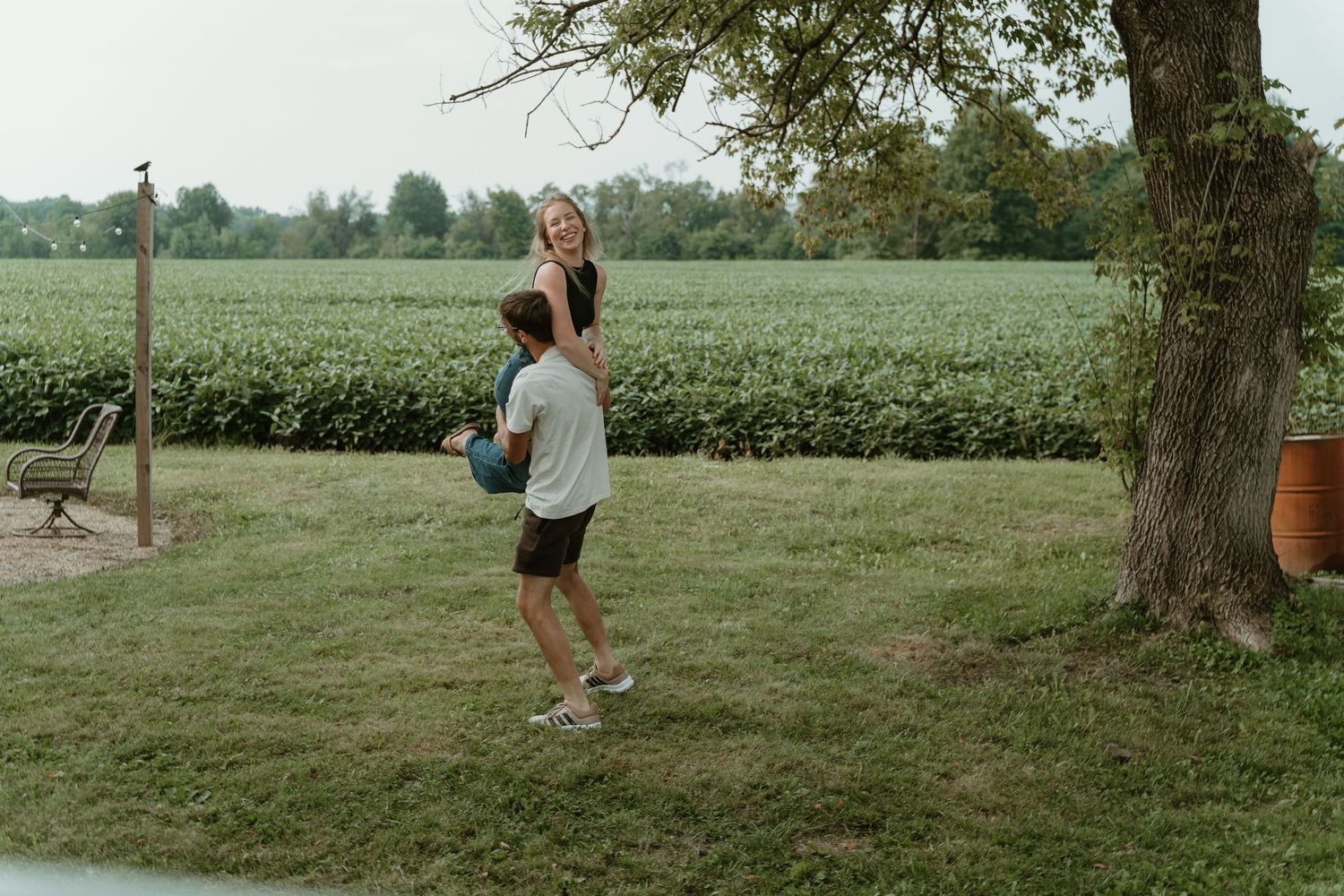 A man lifts a woman outdoors near a tree, with lush green fields in the background.