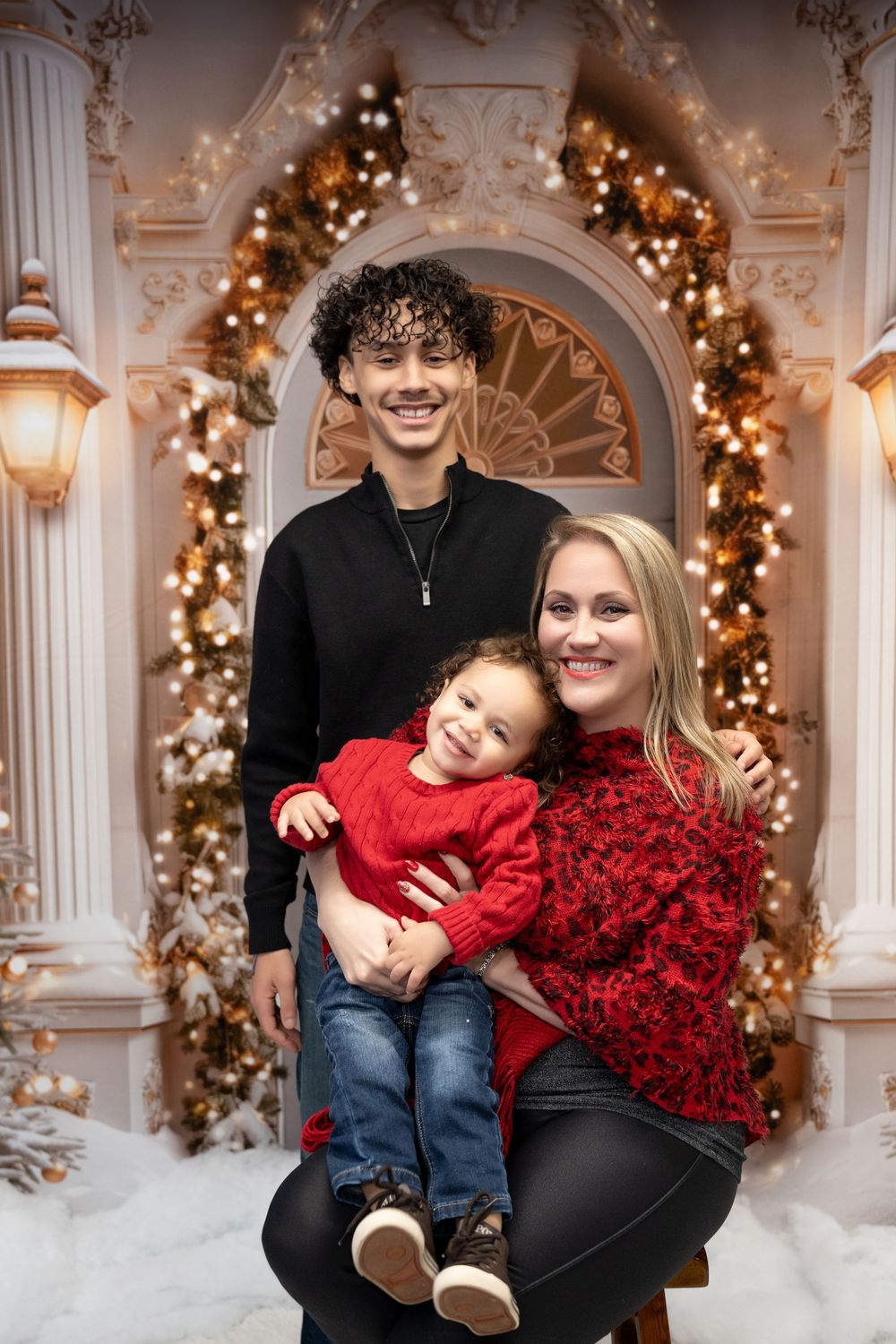 Smiling mother and teenage son pose with laughing toddler in red before elegant Christmas decor.