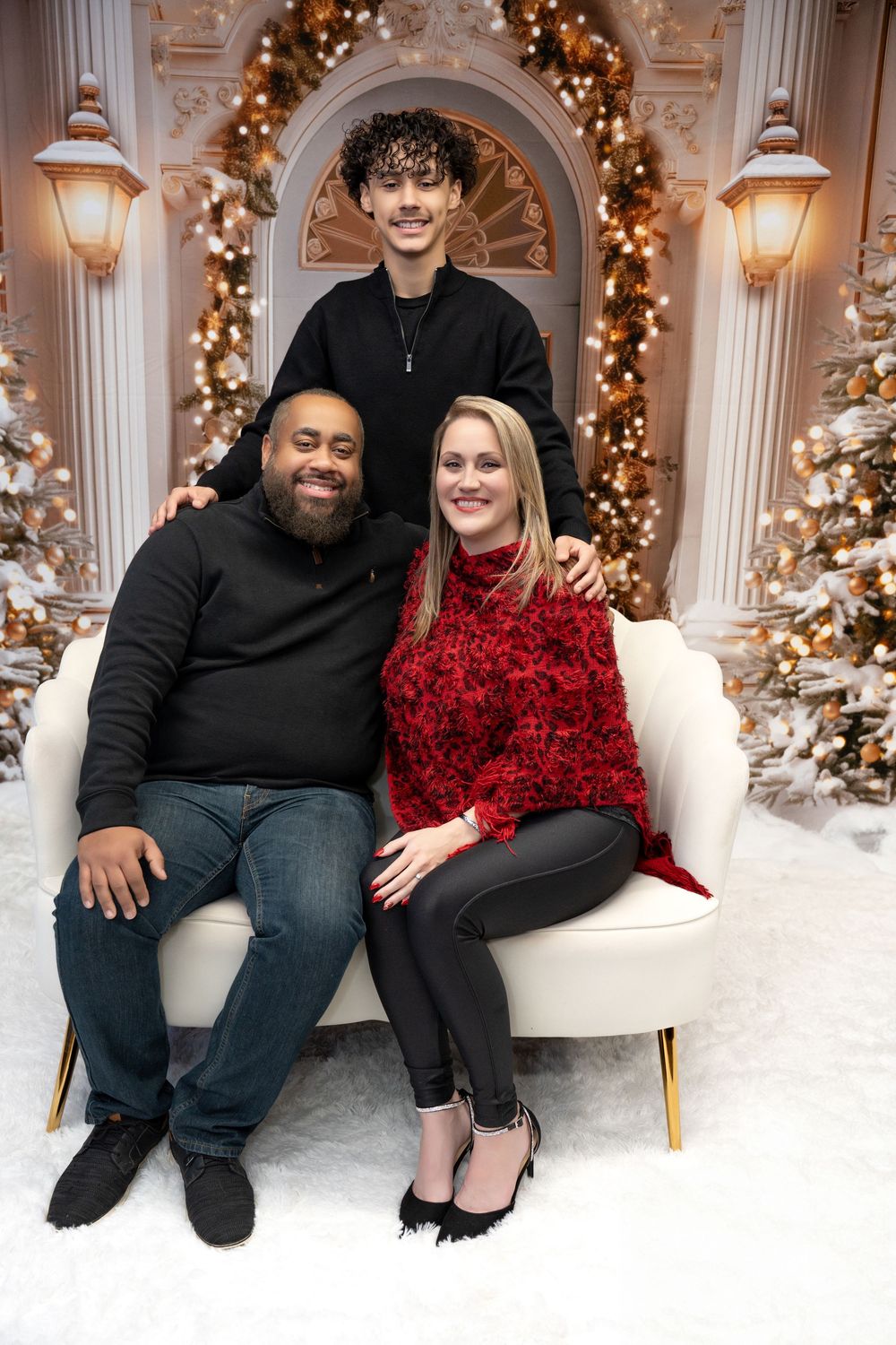 Family of three posing on white sofa with Christmas tree backdrop in holiday portrait.