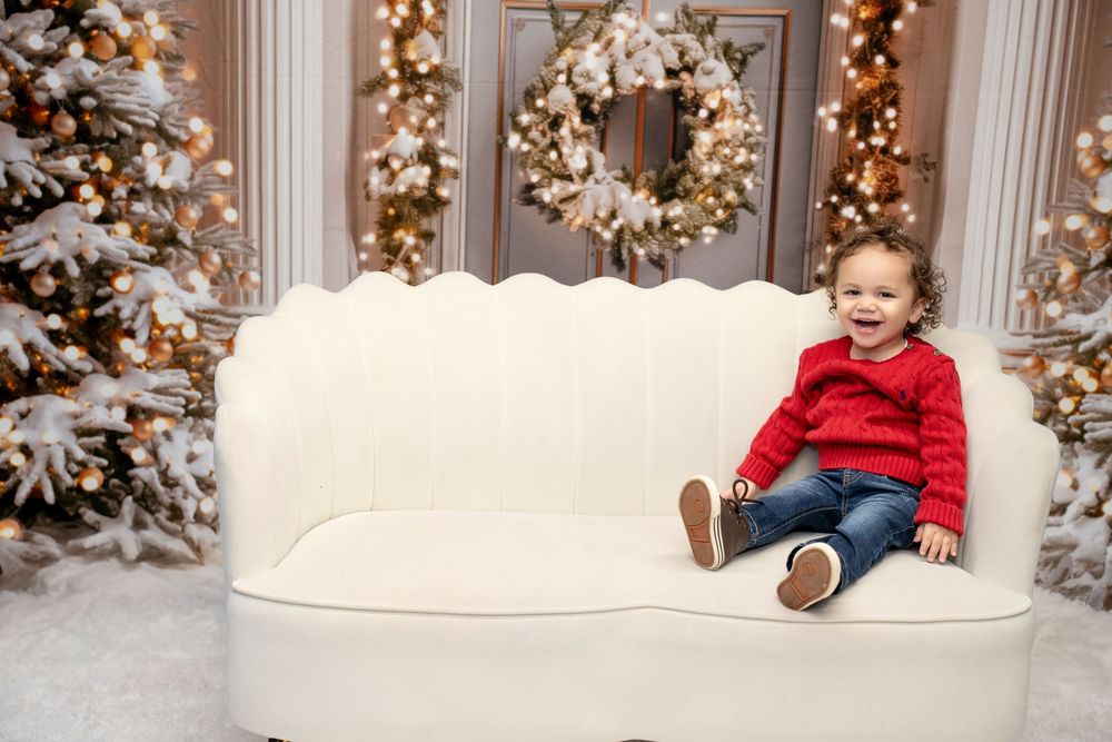 Happy toddler in red sweater sitting on white sofa surrounded by Christmas decorations.