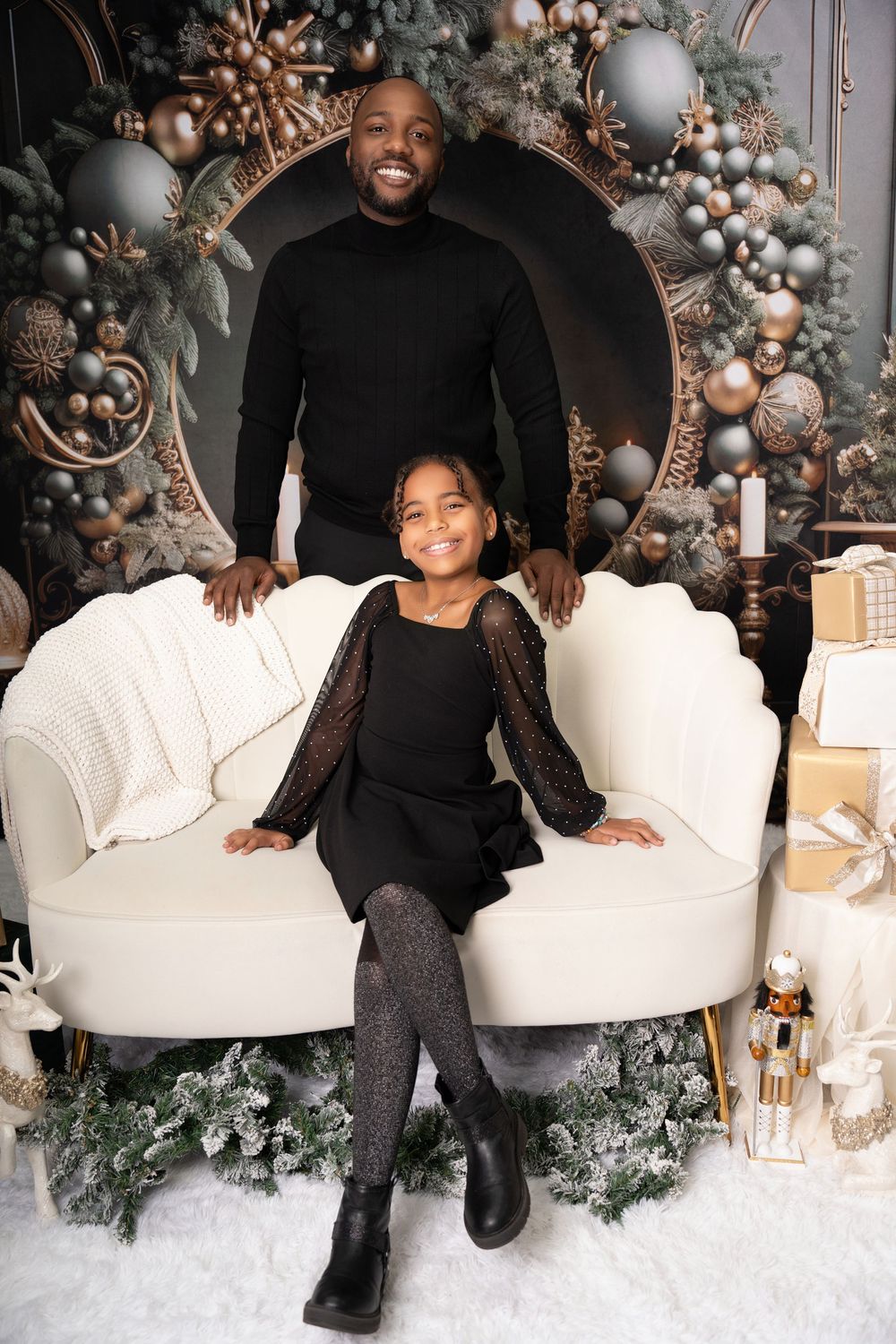 A man and young girl in matching black outfits pose by elegant Christmas decorations with gold ornaments.