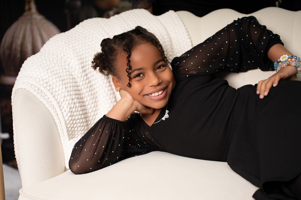 Smiling young girl in black sparkle outfit posing on elegant white sofa.