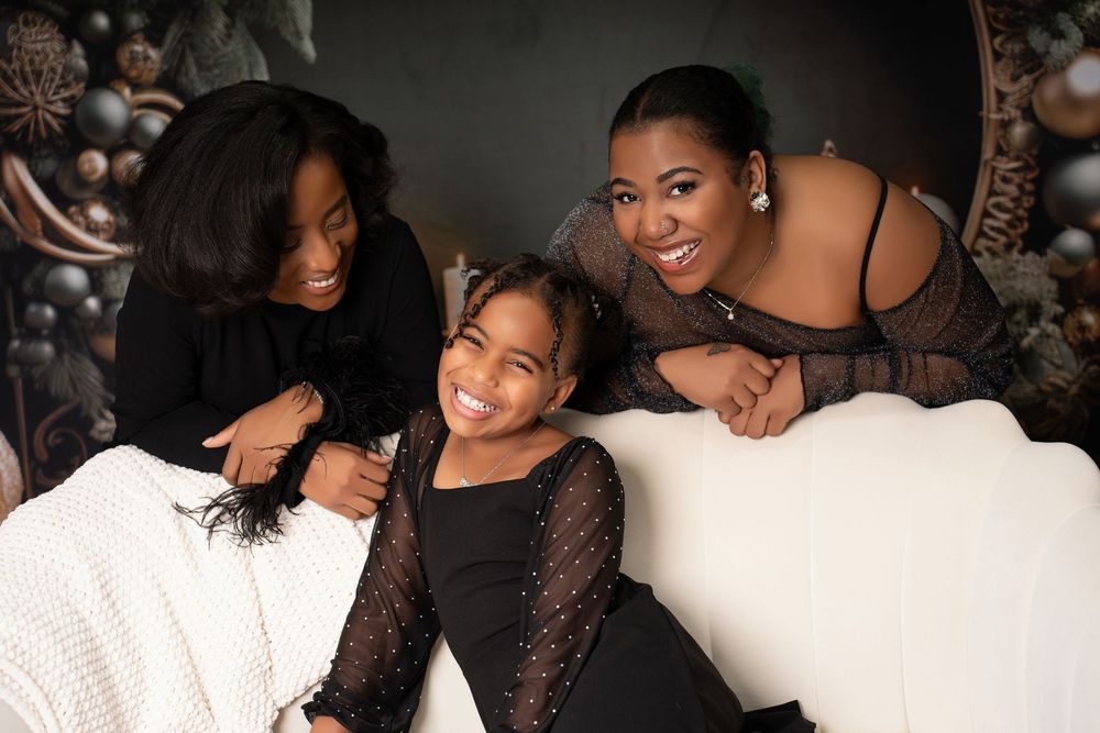 Three smiling women and a young girl laugh together on a white sofa with holiday decor backdrop.