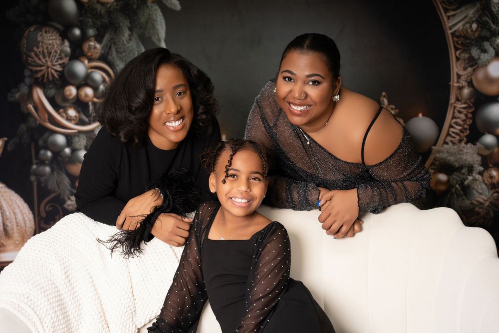 Two women and a young girl in black outfits smile together on a white sofa with Christmas decorations.