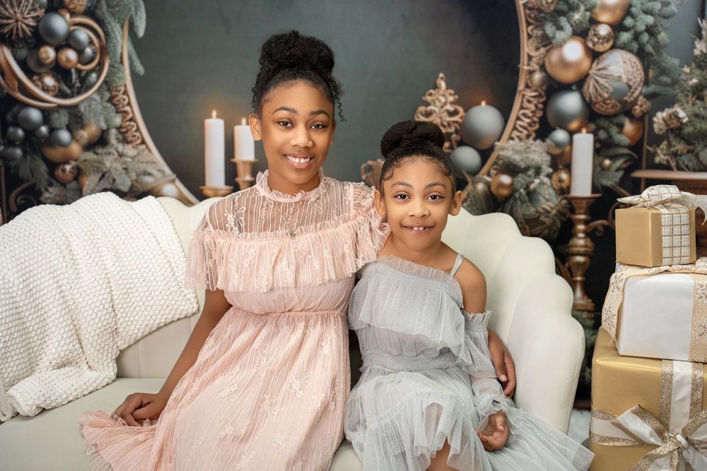 Two girls in elegant dresses sitting together on white sofa with Christmas decor backdrop.