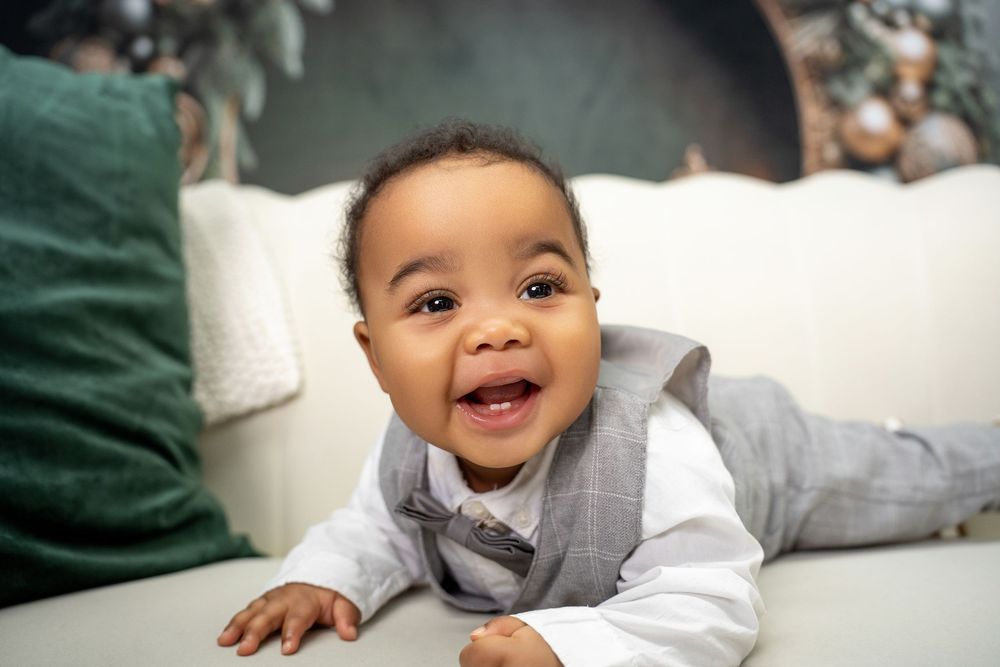 Adorable baby boy in gray vest smiling while doing tummy time on white sofa.