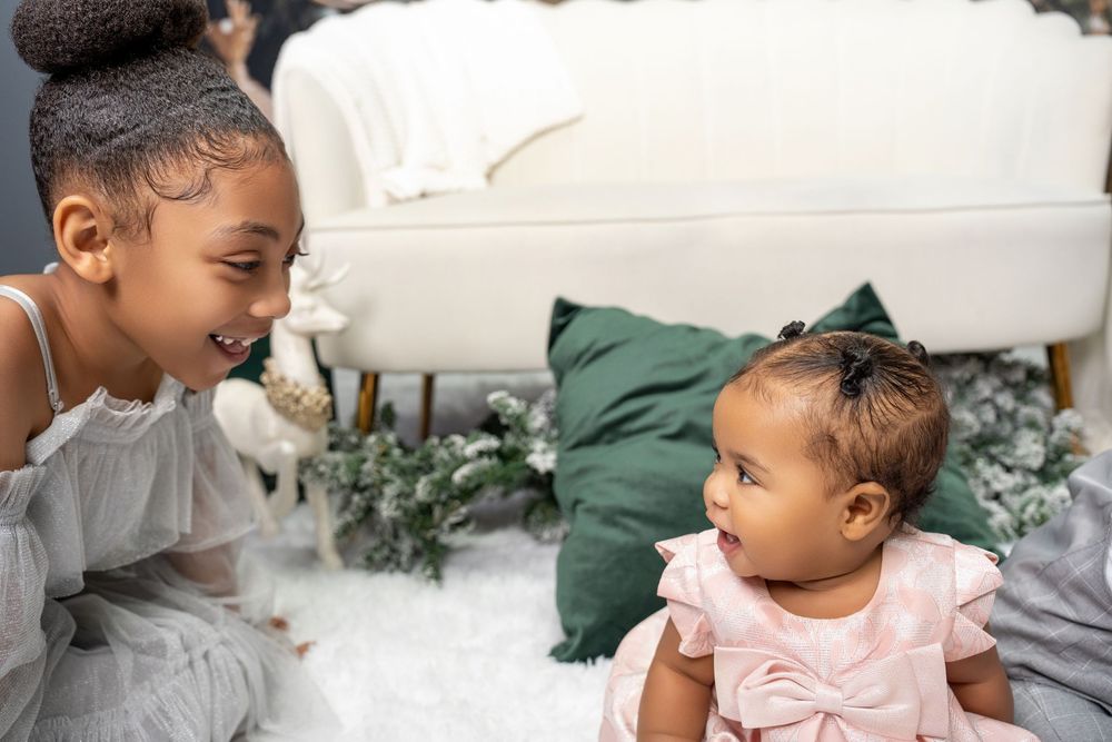 Young girl smiling at baby sister in pink dress during Christmas photo session.