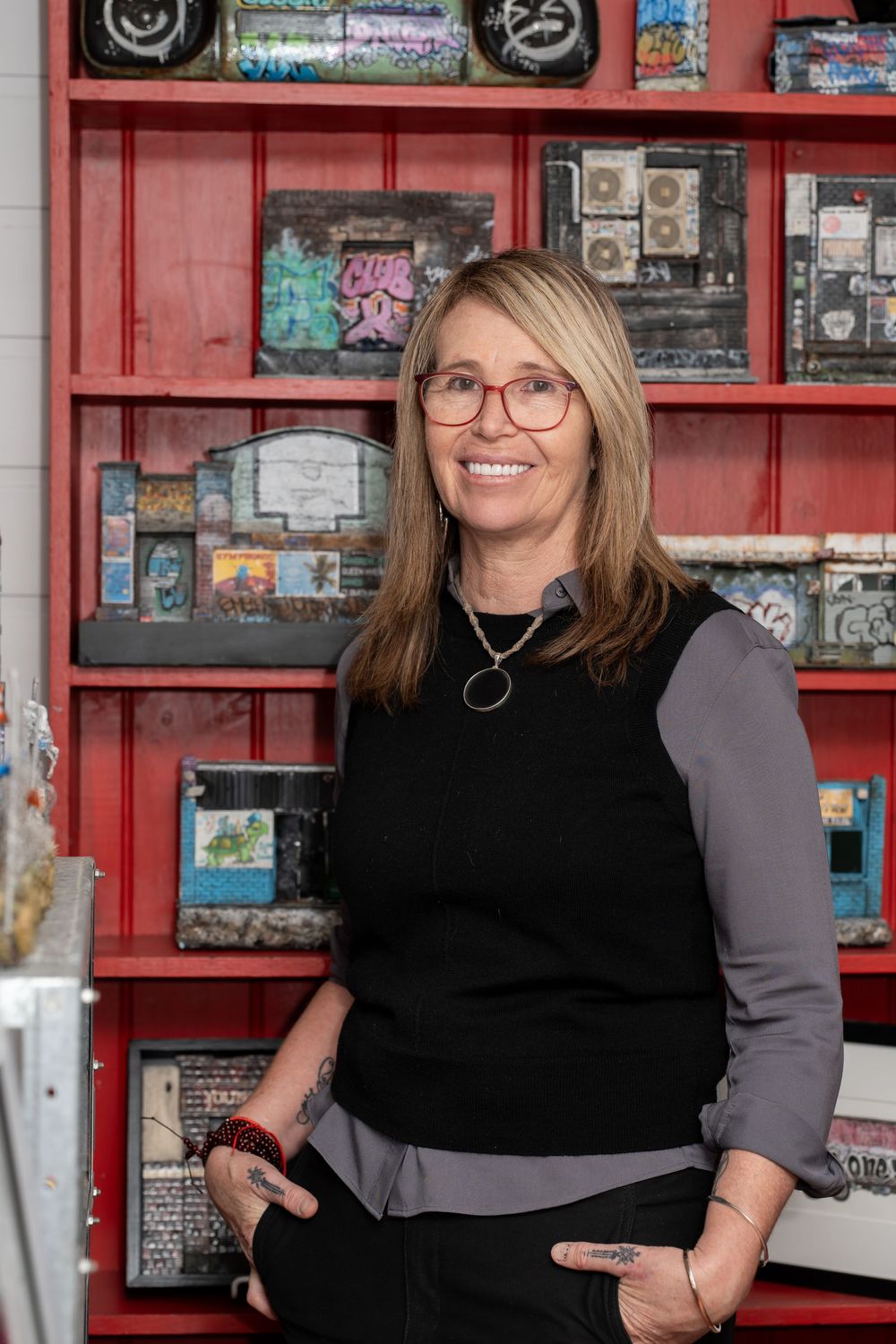 Woman with glasses smiling in art studio, standing before red shelves displaying mixed media artwork.