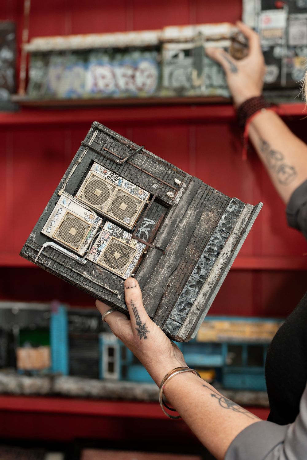 Tattooed hands holding a weathered boombox sculpture in front of graffiti-covered red shelves.