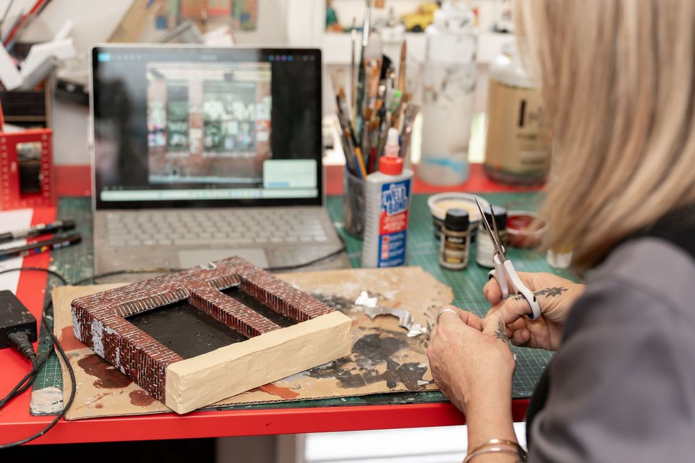 Artist crafting a decorative mosaic frame at a workbench with a laptop, brushes, and craft supplies nearby.
