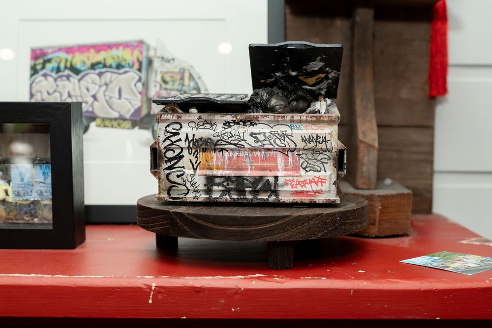Graffiti-covered stacked books on a wooden base displayed on a red table in an art gallery setting.