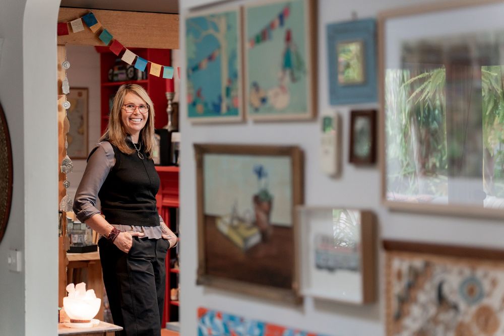 Woman smiling in colorful art studio doorway surrounded by framed paintings and artwork on white walls.