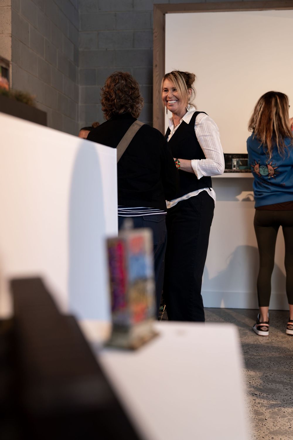Women socializing in a modern gallery space, one smiling in black vest and white shirt.