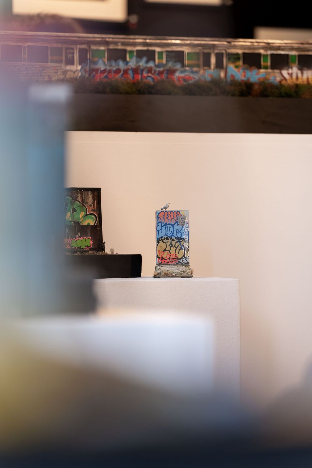 Small graffiti-covered Berlin Wall fragment displayed on white pedestal in modern art gallery with colorful murals in background.