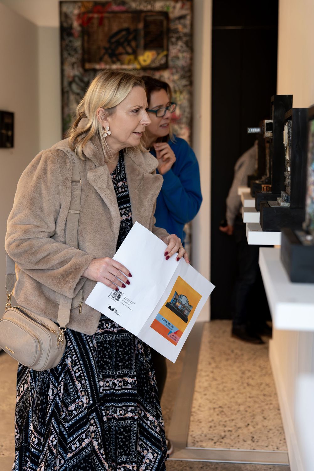 Woman in beige jacket holding art catalog browses paintings at a gallery exhibition with visitors in background.