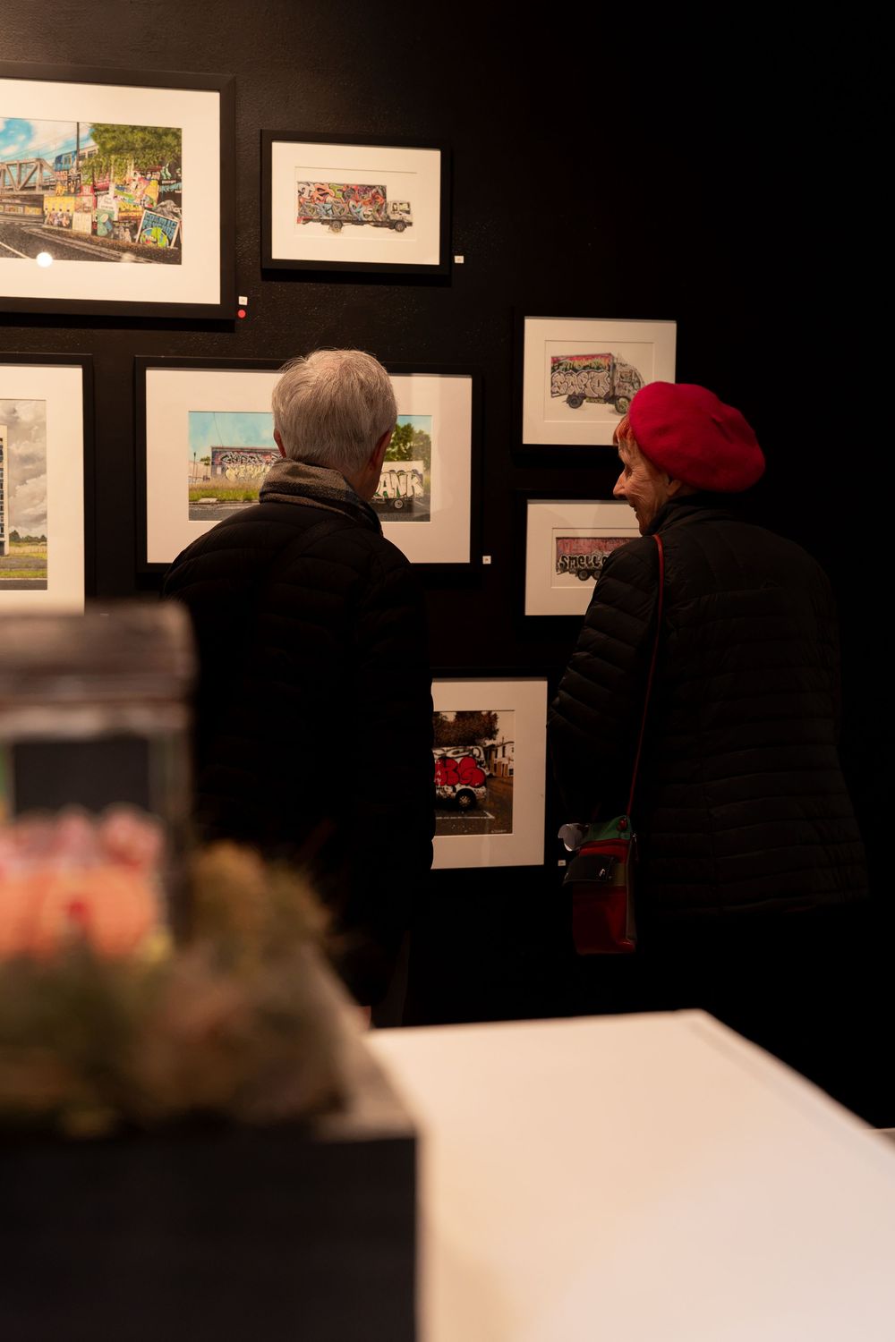 Two people viewing framed artwork prints on a dark gallery wall, one wearing a red beret.