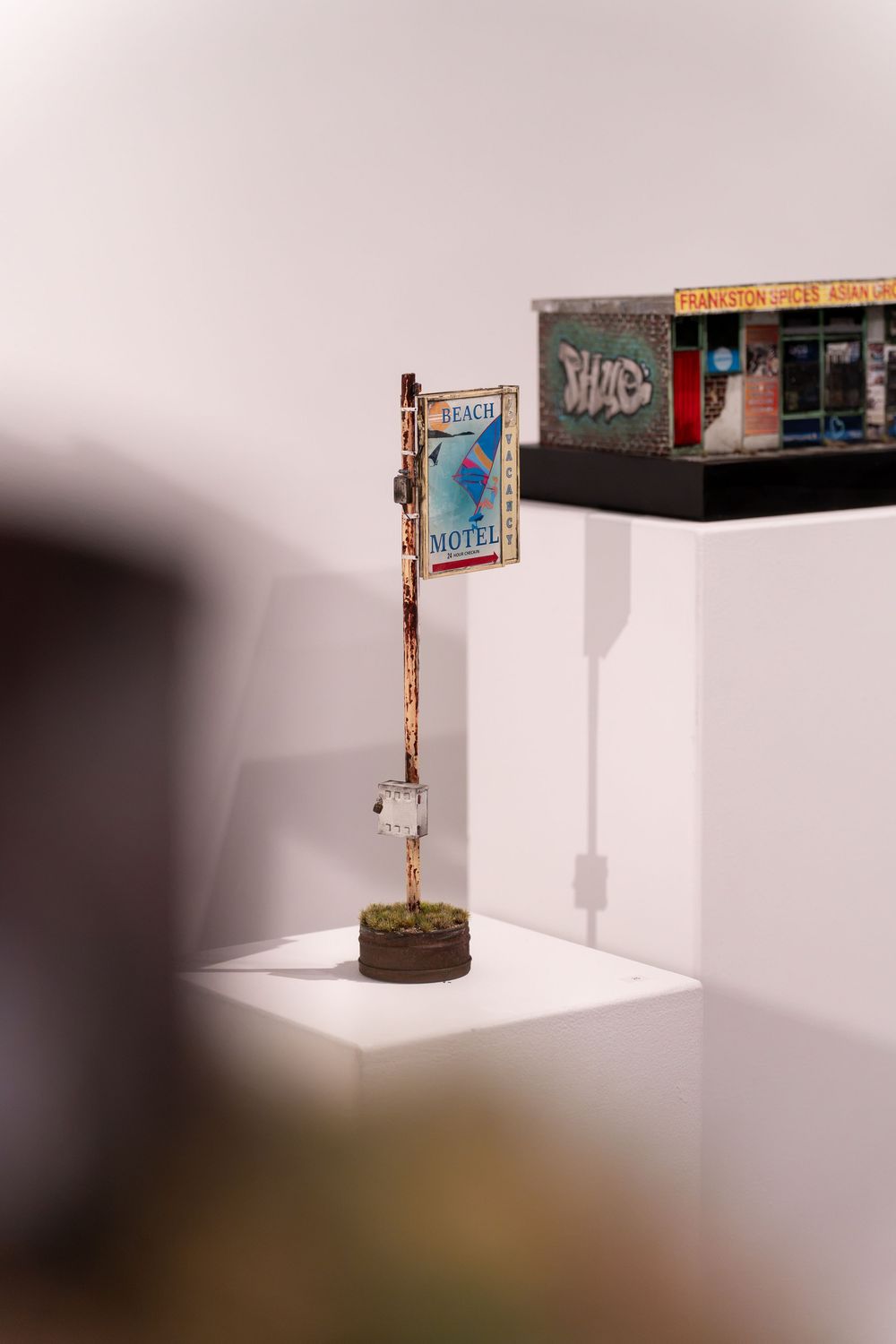 Miniature art installation featuring a rusty Beach Motel sign and a graffiti-covered building model displayed on white shelves.