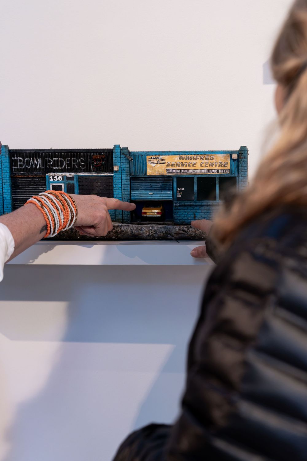 Two people examining a detailed miniature model of a blue urban building at an art gallery exhibition.