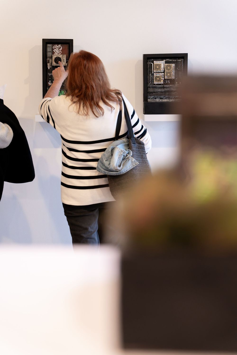 Woman in striped jacket viewing framed artwork on white gallery wall while carrying a gray shoulder bag.