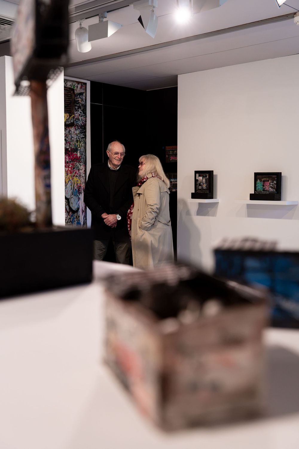 Two visitors converse in a contemporary art gallery with framed artwork on white walls and a sculptural piece in the foreground.