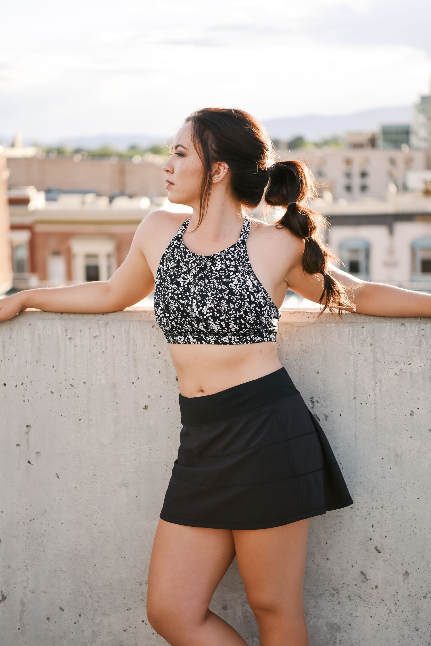 Woman in black skirt and patterned crop top posing on urban rooftop with city skyline background.