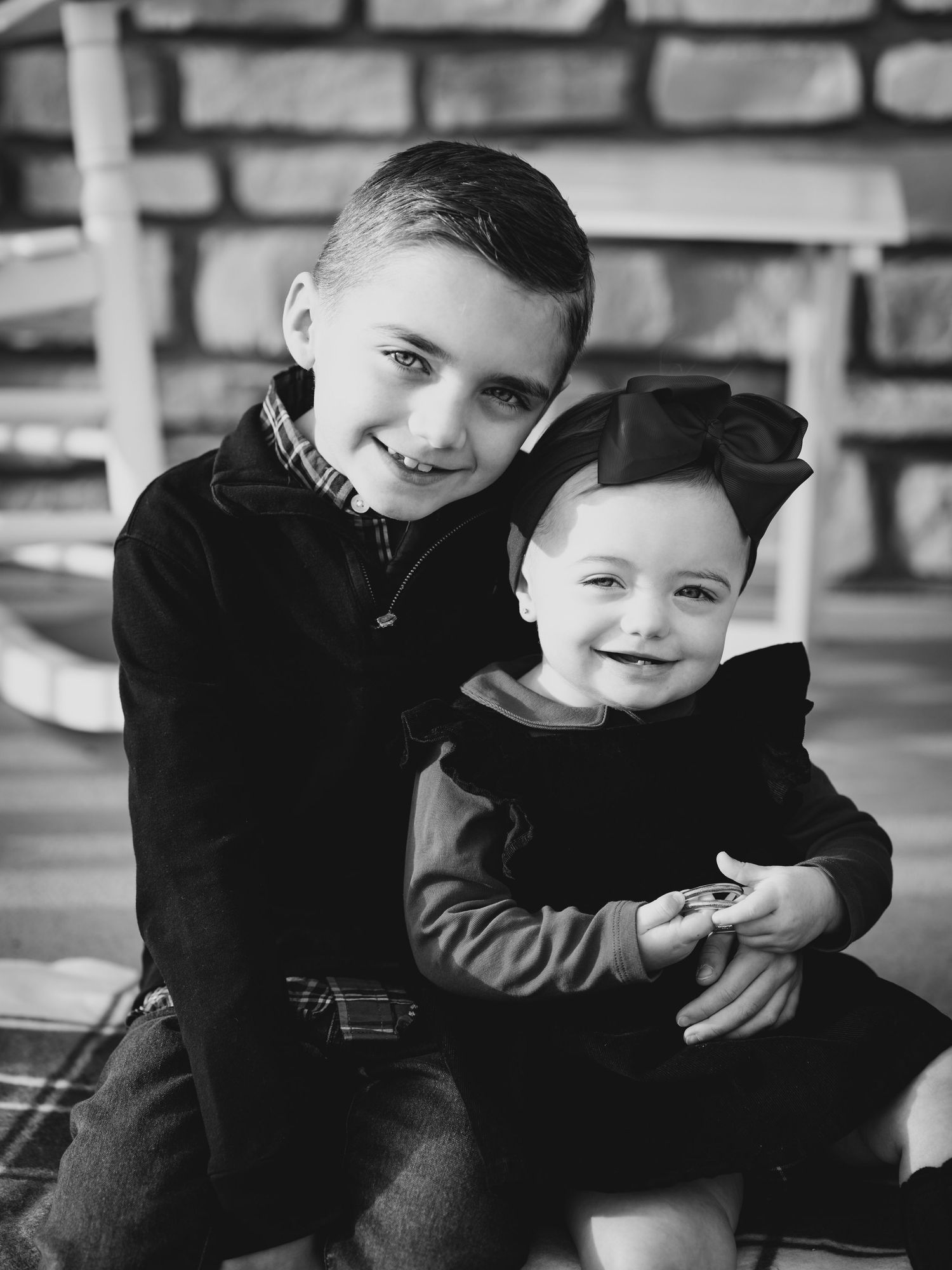 Black and white photo of two smiling siblings, older boy hugging younger toddler girl wearing a bow, seated outdoors.