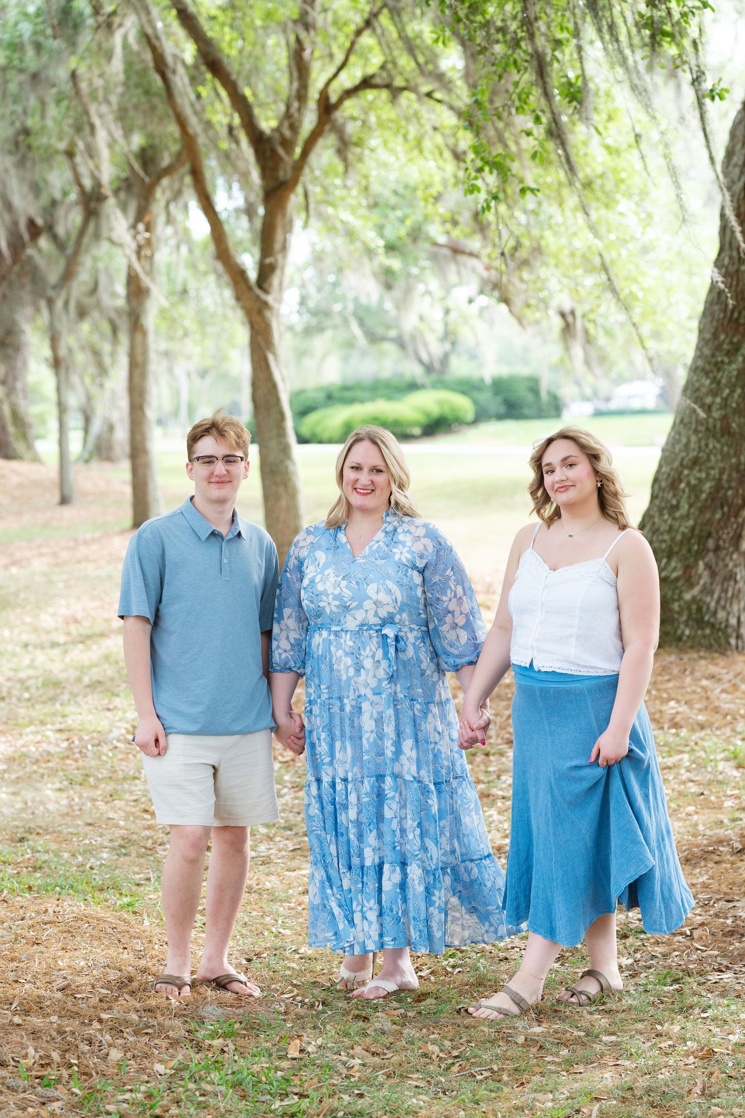 Family of three in coordinated blue outfits posing beneath oak trees draped in Spanish moss.