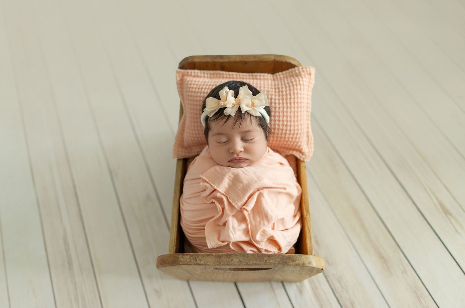 Newborn baby sleeping peacefully in a tiny wooden bed, wrapped in peach fabric with a floral headband.