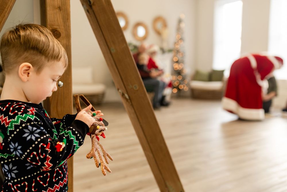 A toddler in a Christmas sweater holds a reindeer ornament near a wooden easel with Santa Claus visible in the background.