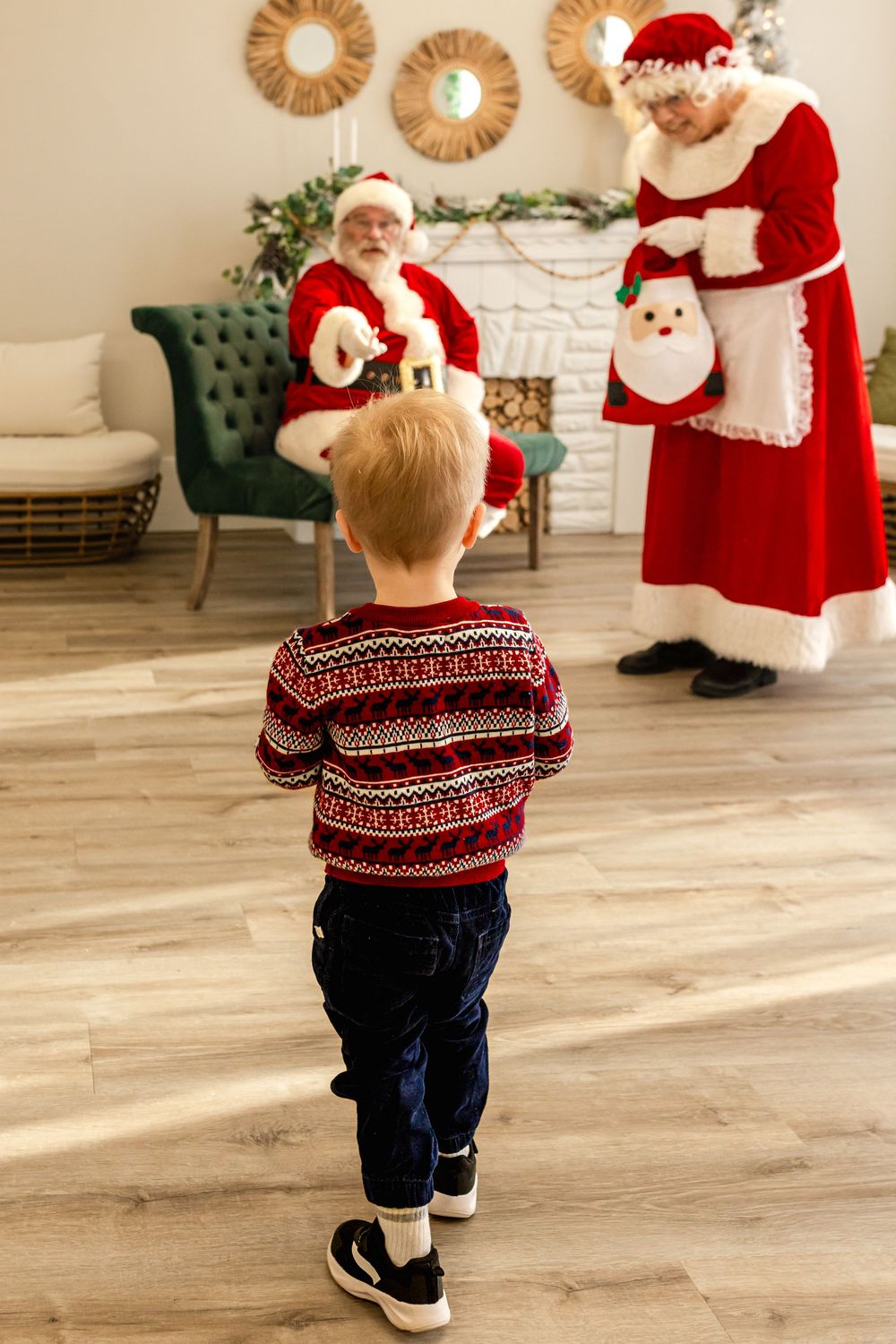 A young boy in a festive sweater walks toward Santa Claus and Mrs. Claus in a decorated holiday room.