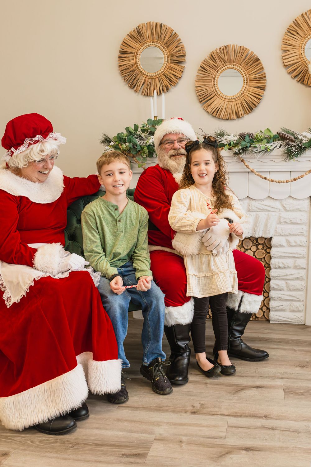 Two children pose with Santa and Mrs. Claus in front of a festive fireplace decorated with garland and mirrors.