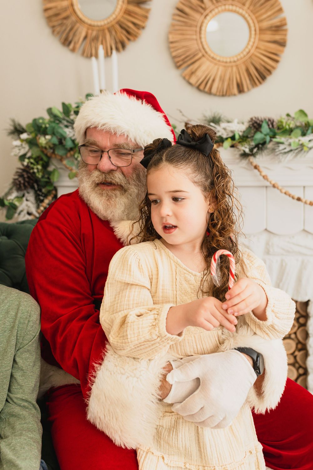 A young girl sits on Santa Claus's lap holding a candy cane near a decorated fireplace mantel.