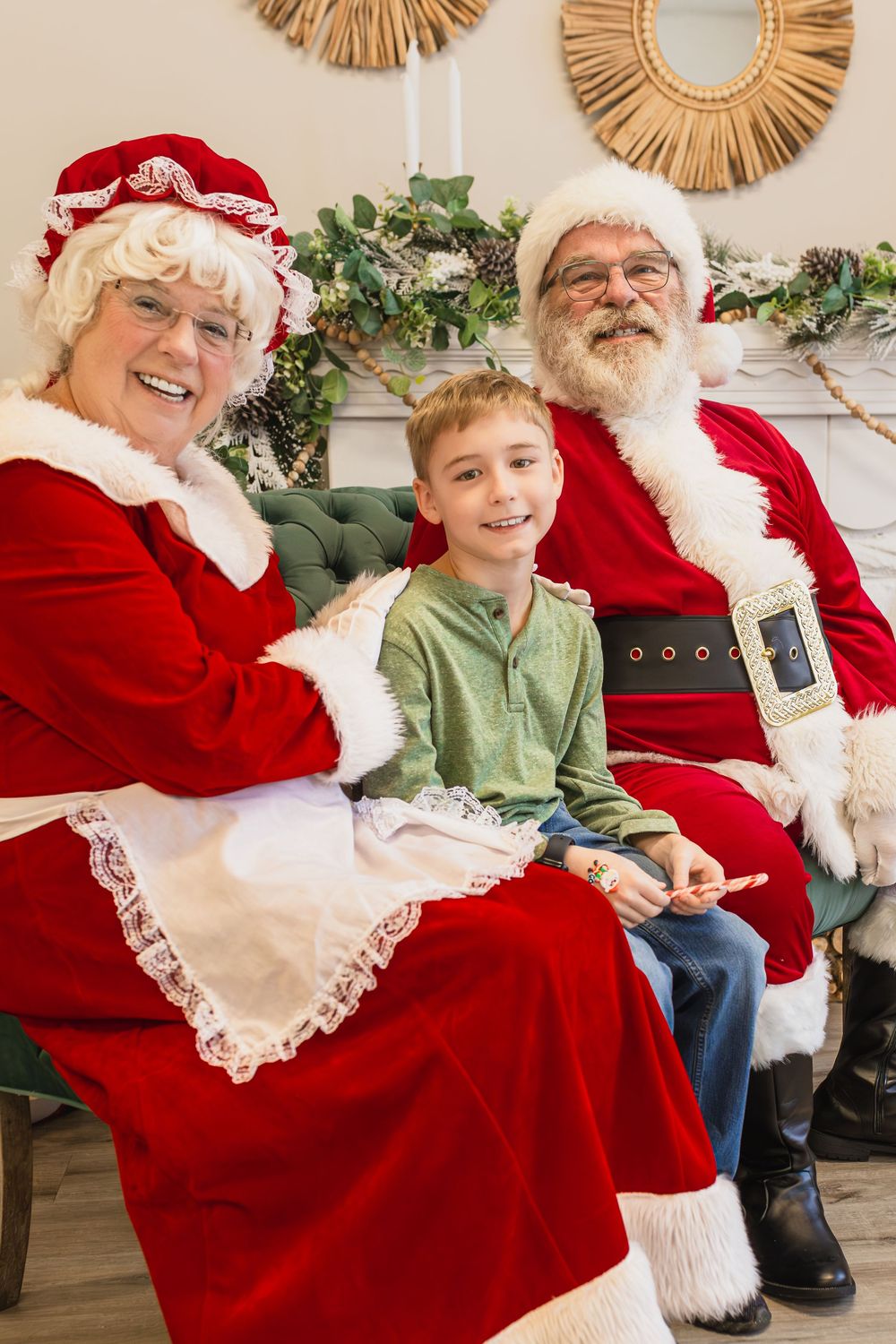 A young boy sits smiling between Santa Claus and Mrs. Claus in festive red costumes near a decorated fireplace.