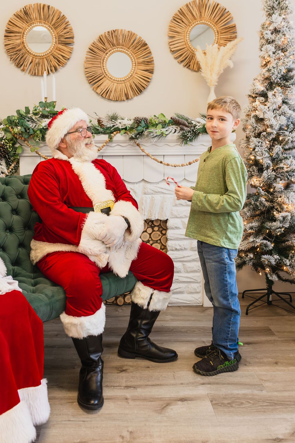 A boy stands talking to Santa Claus near a decorated Christmas tree and fireplace in a festive holiday setting.