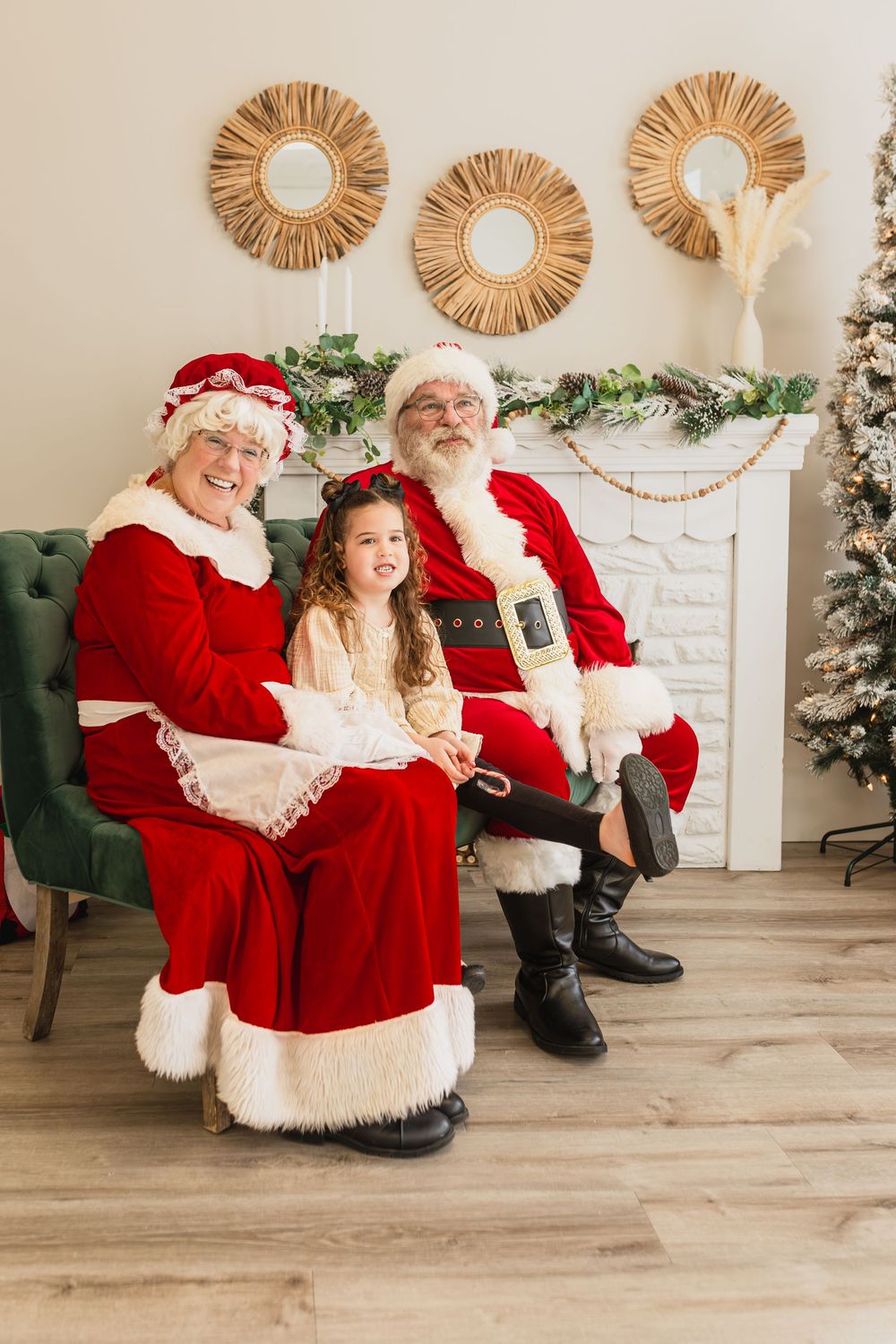 A young girl sits between Santa Claus and Mrs. Claus in a festive Christmas setting with a decorated fireplace.