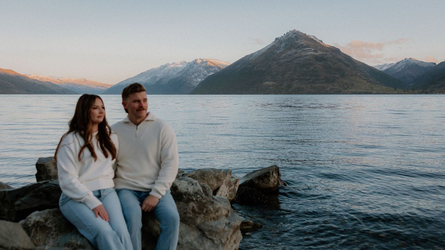 Couple in matching white sweaters and jeans sit together on lakeside rocks with snow-capped mountains glowing at sunset behind them.