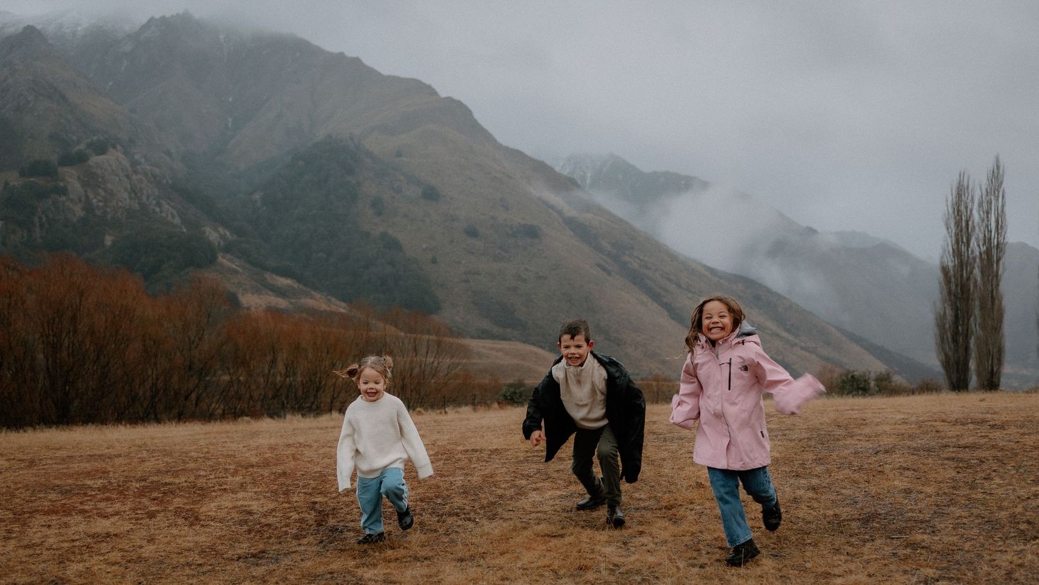 Three children run through a golden grassy field with misty mountains and bare trees rising in the moody background.