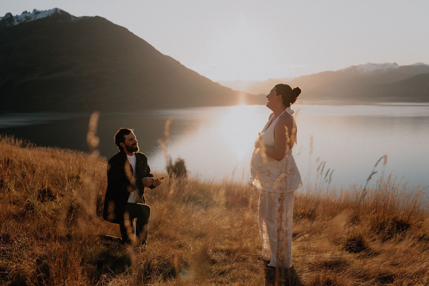 Man kneeling on one knee proposes to a woman in a flowing white dress on a golden grassy hillside beside a mountain lake at sunset.