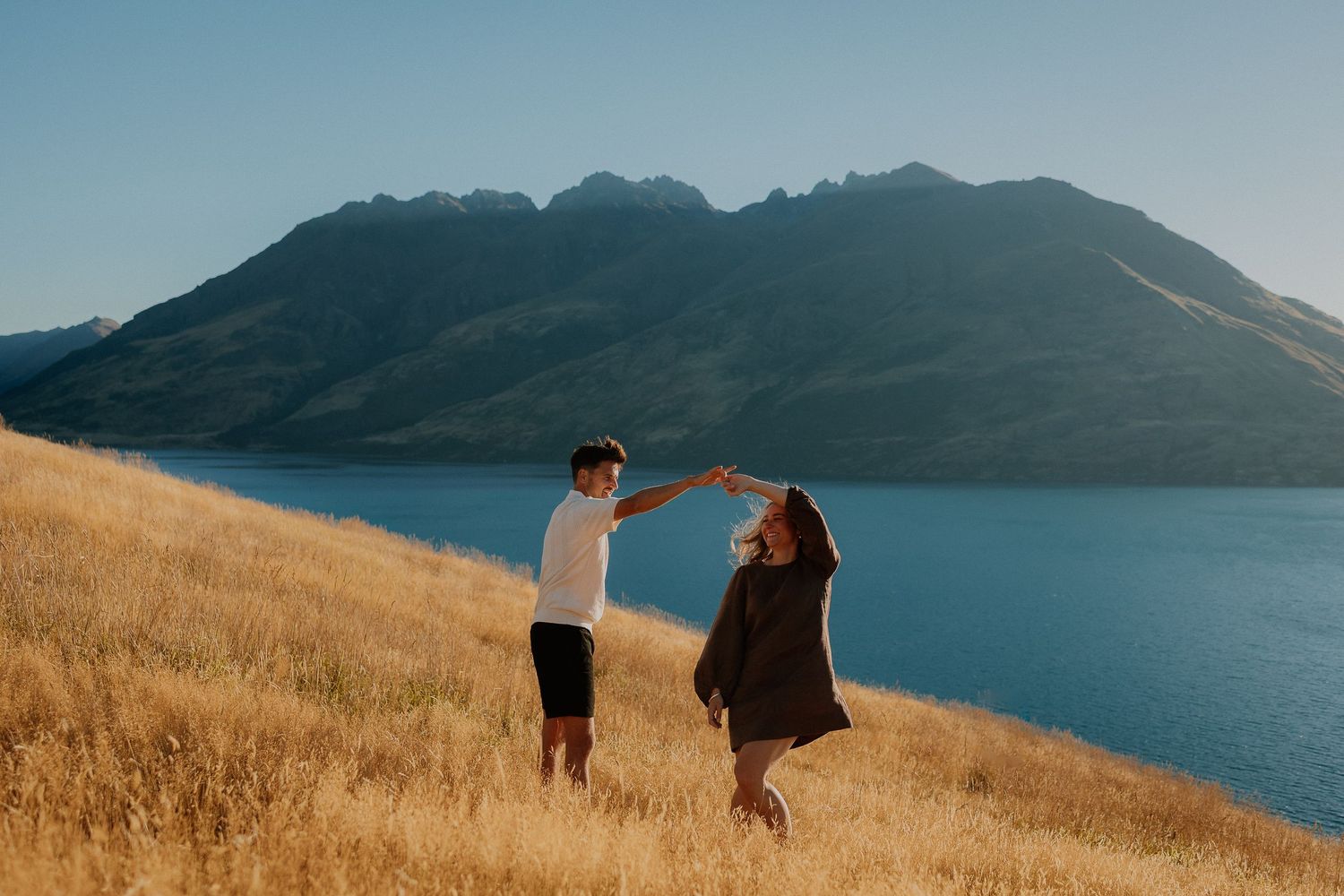 Couple dancing hand in hand on a golden grassy hillside overlooking a calm blue lake and rugged mountains at sunset.
