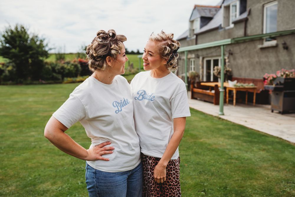 Two women in matching white t-shirts with hair in curlers smile at each other on a sunny garden lawn outside a stone country house.