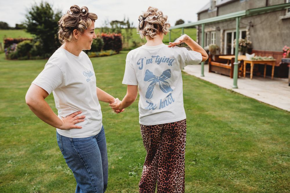 Two women with pinned-up hair hold hands and walk across a sunny lawn, one in a white tee and jeans, the other in leopard print pants.