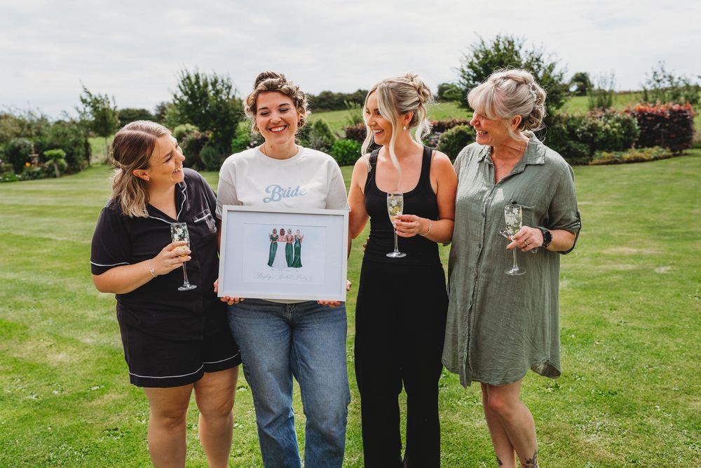 Four smiling women hold champagne glasses and a framed illustration while posing together on a sunny green lawn with shrubs behind them.